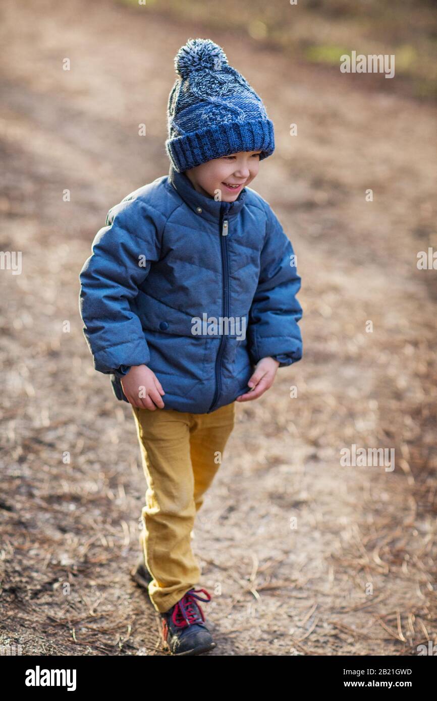 Piccolo ragazzo carino con giacca calda blu e cappello lavorato a maglia sorridendo contro il parco sfocato a basse temperature Foto Stock