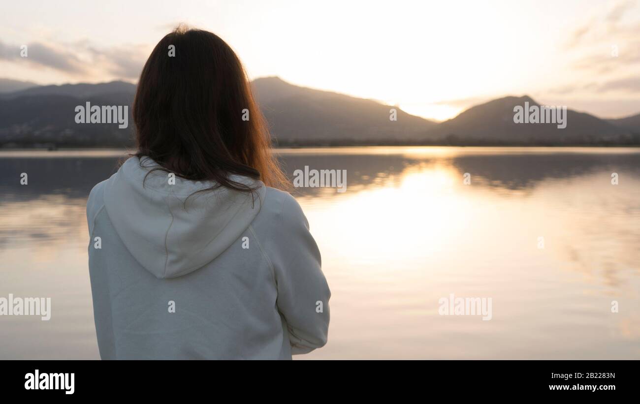 Giovane donna penosa con capelli lunghi visto da dietro guarda il lago al tramonto Foto Stock