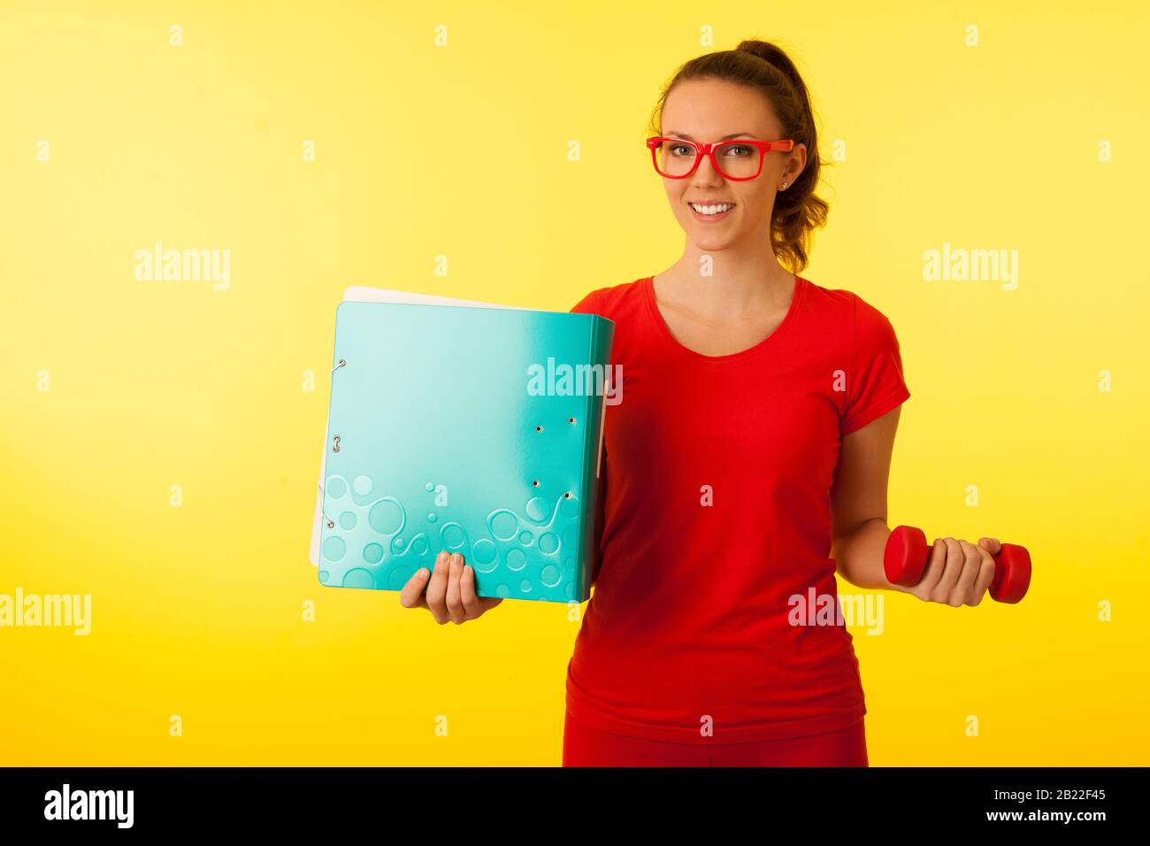 cute giovane felice caucasica donna in t shirt rossa su sfondo giallo vibrante gesto potere di conoscenza hodling un dumbbell e grande cartella Foto Stock