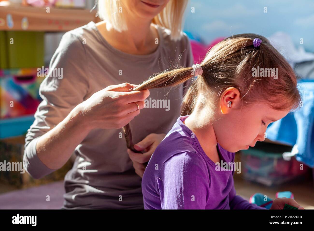 La madre styling i suoi capelli delle figliuole, tenendolo fare i suoi ponytails. Mamma e figlia chiudono momenti intimi. Bambini, capelli bambini, luce solare, bambini Foto Stock