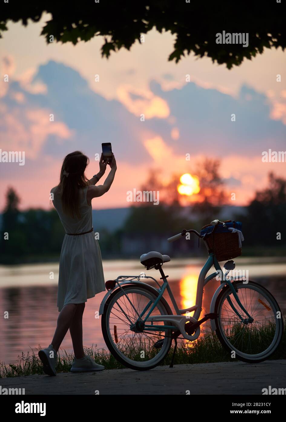 Lady vicino a una moto retrò scatta una foto al tramonto su un telefono cellulare. Una ragazza vestita di un abito leggero con un arco sulla testa sta cercando di catturare il sole durante un bel tramonto vicino al lago Foto Stock