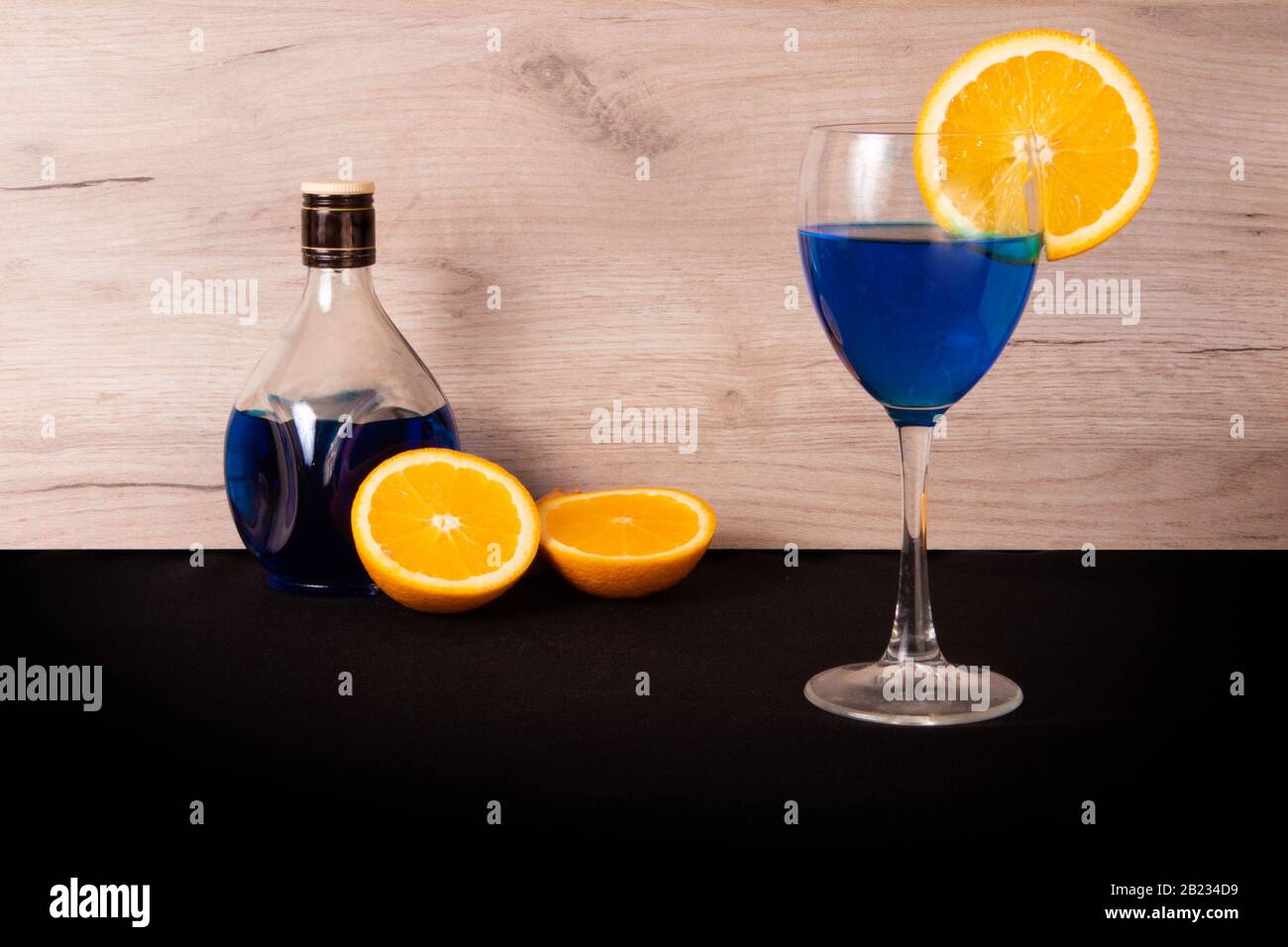 Bicchiere con bevanda blu e fetta di arancia succosa sul lato, bottiglia e metà arancioni rovesciate dietro Foto Stock