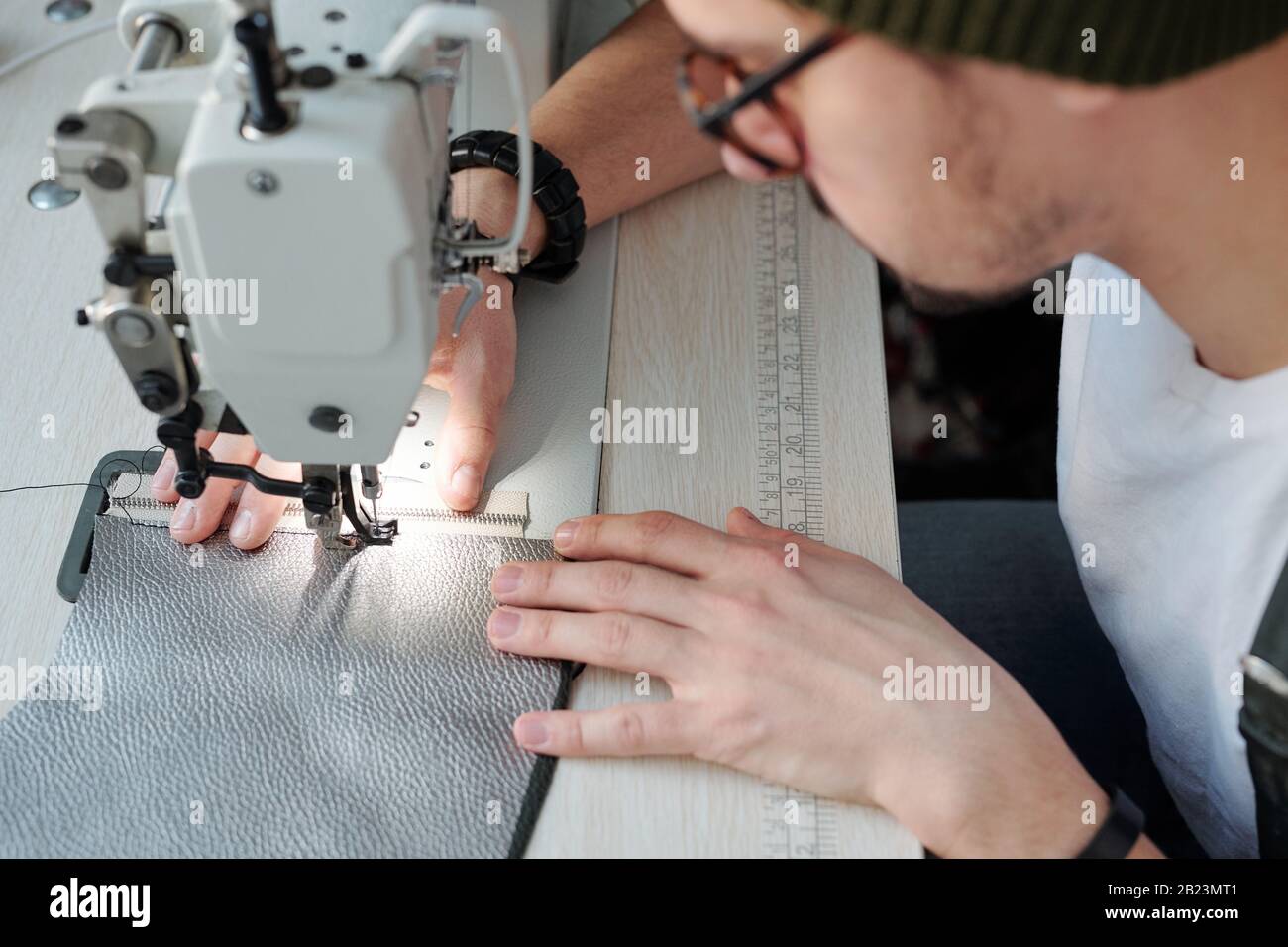 Mani di giovani pellettieri che cucono zip e pezzi di cuoio insieme mentre piegano sopra macchina elettrica in officina Foto Stock