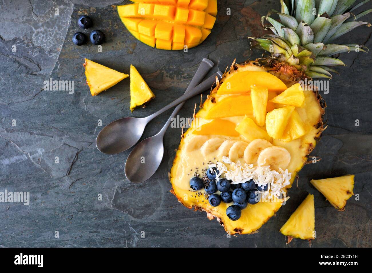 Ciotola sana di frullato in un ananas con cocco, banane, mango e mirtilli. Vista dall'alto su uno sfondo scuro di ardesia. Spazio di copia. Foto Stock