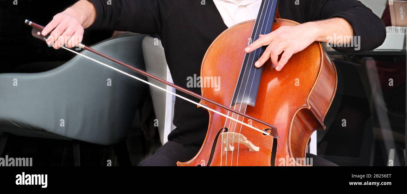 Un banner in formato computer mostra un primo piano delle mani di un uomo che gioca un violoncello in un ambiente esterno. Foto Stock