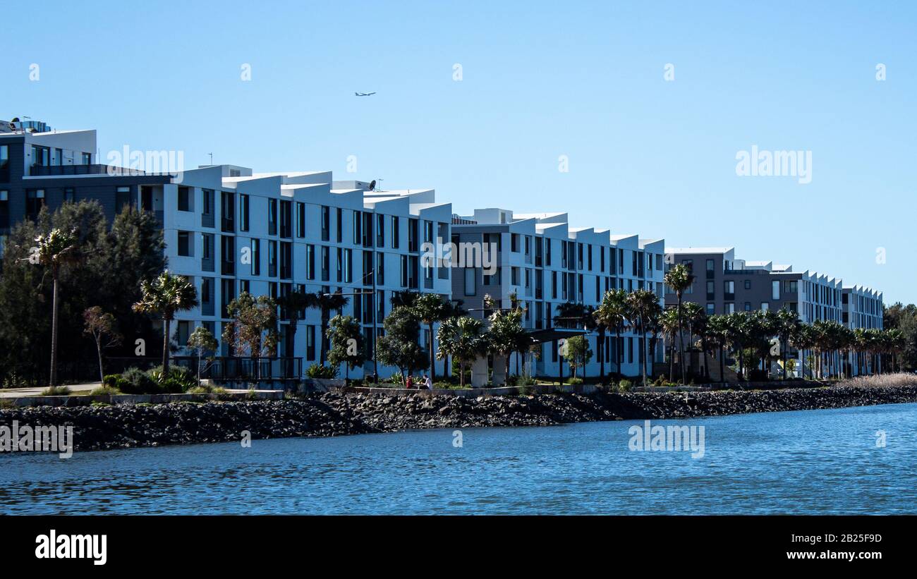 Condomini appartamento fronte mare nella comunità suburbana sulla riva del fiume con passerella alberata, cielo blu sullo sfondo Foto Stock