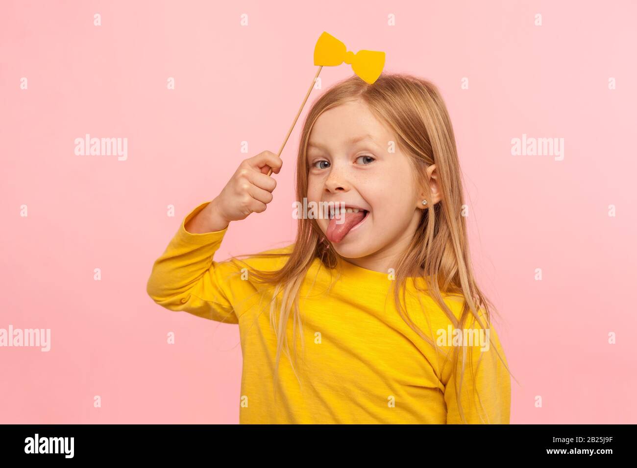 Ritratto di giocoso adorabile bambina di zenzero che tiene arco di carta e che si attacca la lingua, bambino naughty che si diverte, indossando accessorio masquerade. IND Foto Stock