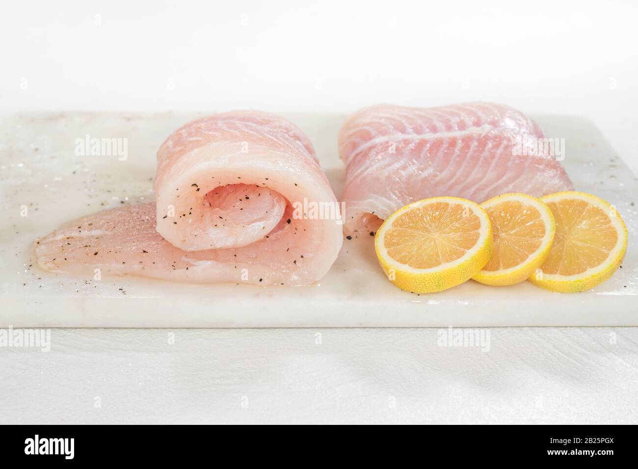 Filetto di pesce fresco crudo. Filetto di spigola e fette di limone rosa si chiudono su un tagliere di marmo su sfondo bianco, vista frontale Foto Stock