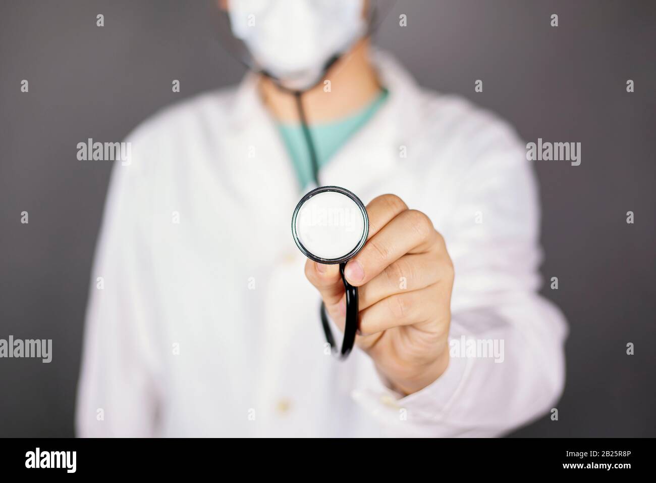 medico in un abito medico bianco con una maschera usa e getta e uno statoscopo nelle sue mani in piedi su uno sfondo scuro. Foto Stock