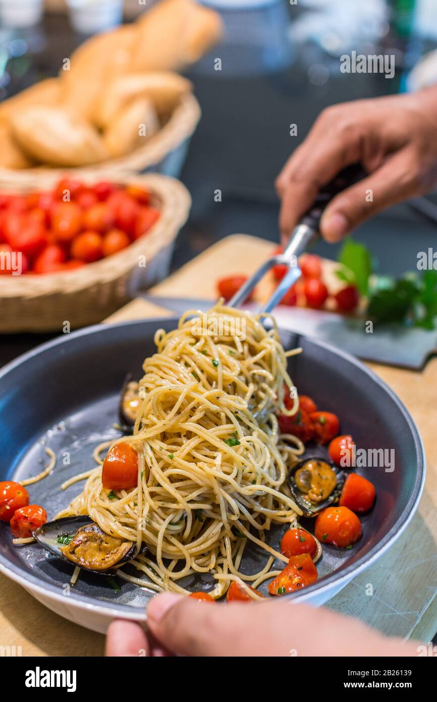 Primo piano di uno chef che prepara spaghetti Vongole con pomodori e cozze roma in una padella - con un coltello e tagliere con pomodori e pane sul retro Foto Stock