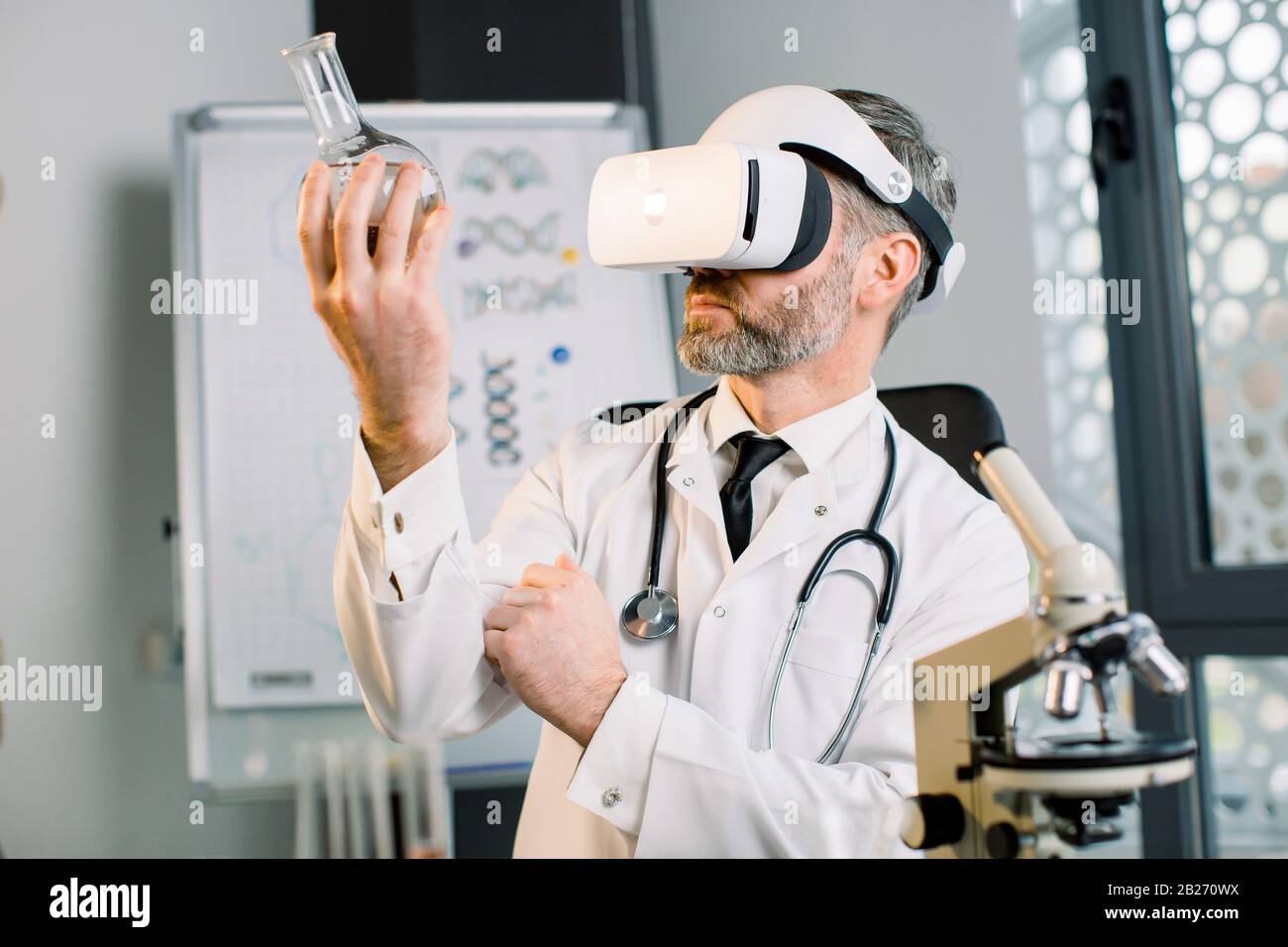 Uomo scienziato maturo in moderno laboratorio luminoso, utilizzando occhiali di realtà virtuale per la ricerca, guardando il pallone di vetro con nuova sostanza o droga. Vr Foto Stock
