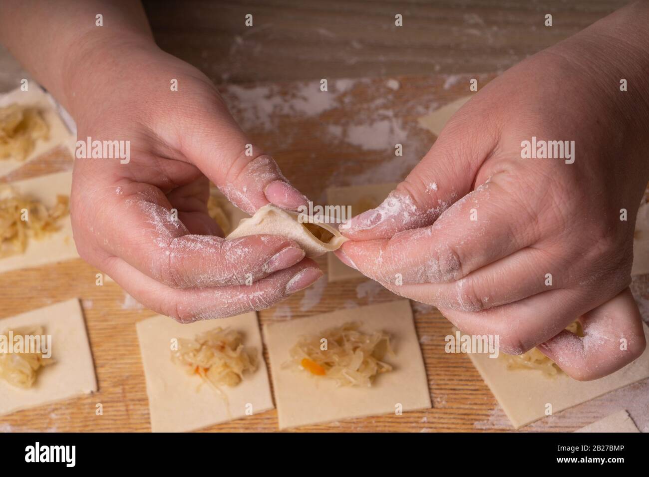 Una donna scolpisce gnocchi e ravioli da quadrati di pasta e cavolo. Pannello di taglio compensato, setaccio di farina di legno e perno di laminazione di legno - attrezzi per Foto Stock
