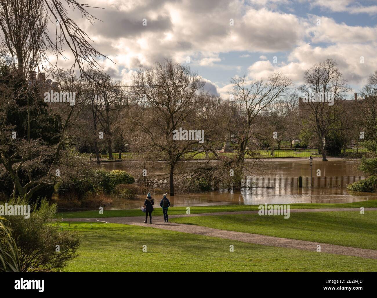 Inverno nei giardini pubblici di York. Un sentiero si estende verso un fiume inondato con un albero spezzato e due giovani guardano. Foto Stock