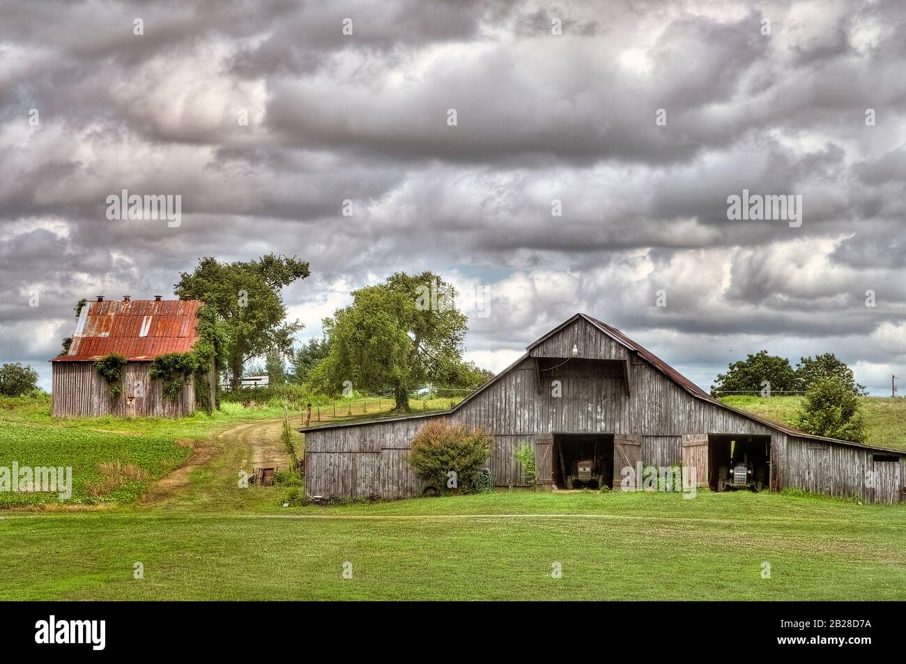 Due barre usurate e stagne si siedono con le loro porte aperte e i trattori a riposo sotto un cielo nuvoloso e dinamico Foto Stock