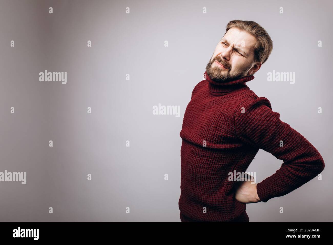 Uomo bearded in maglia calda maglione in piedi in studio con espressione dolorosa e toccando la schiena a causa di forte dolore. Persona matura sensazione spi Foto Stock