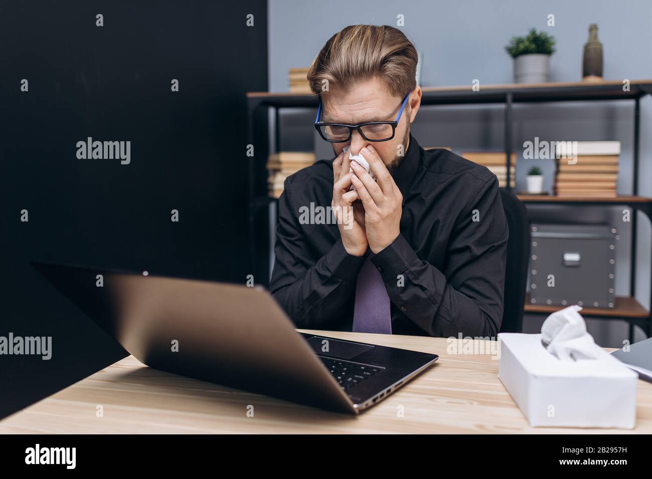 Uomo d'affari stanco e malato in abbigliamento formale e occhiali che soffiano il naso in tovagliolo mentre si lavora su un portatile presso il centro ufficio. Concetto di malattia e. Foto Stock