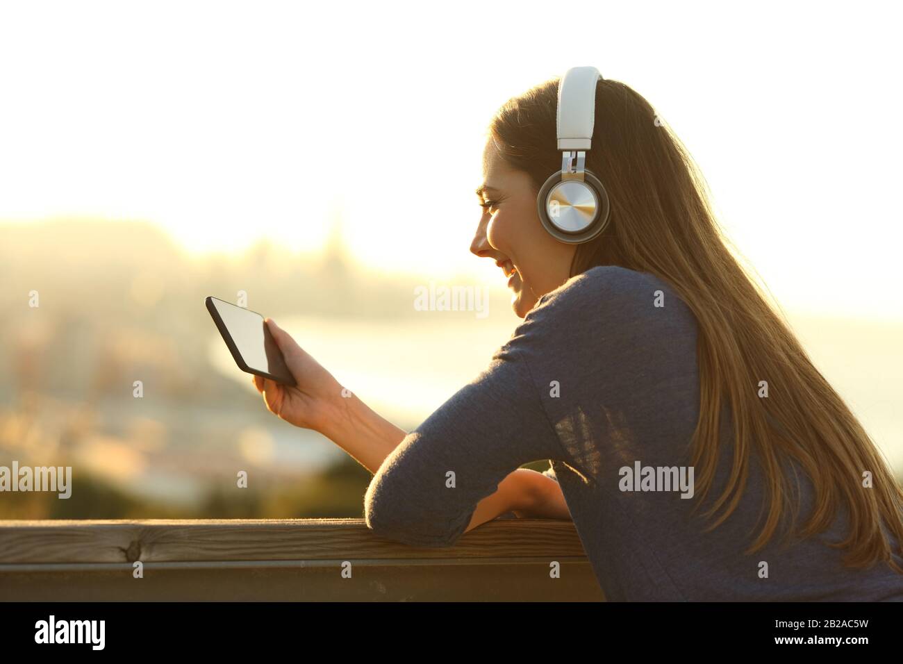 Donna felice che indossa le cuffie per guardare video su smartphone in un balcone con vista sulla costa al tramonto Foto Stock