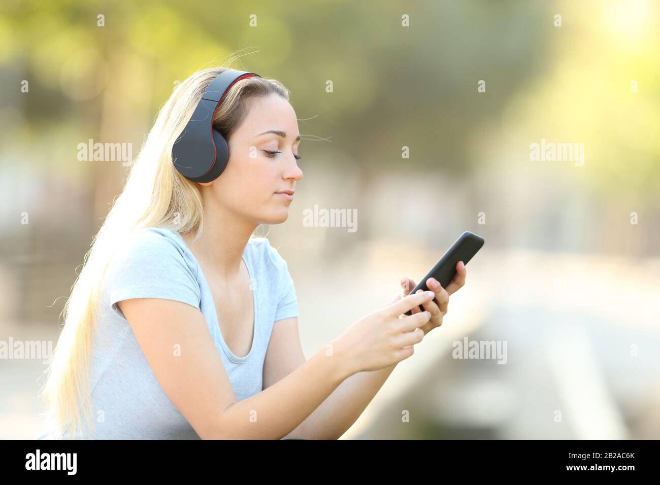 Ragazza giovane bionda che ascolta la musica sul suo telefono con le cuffie in un parco Foto Stock