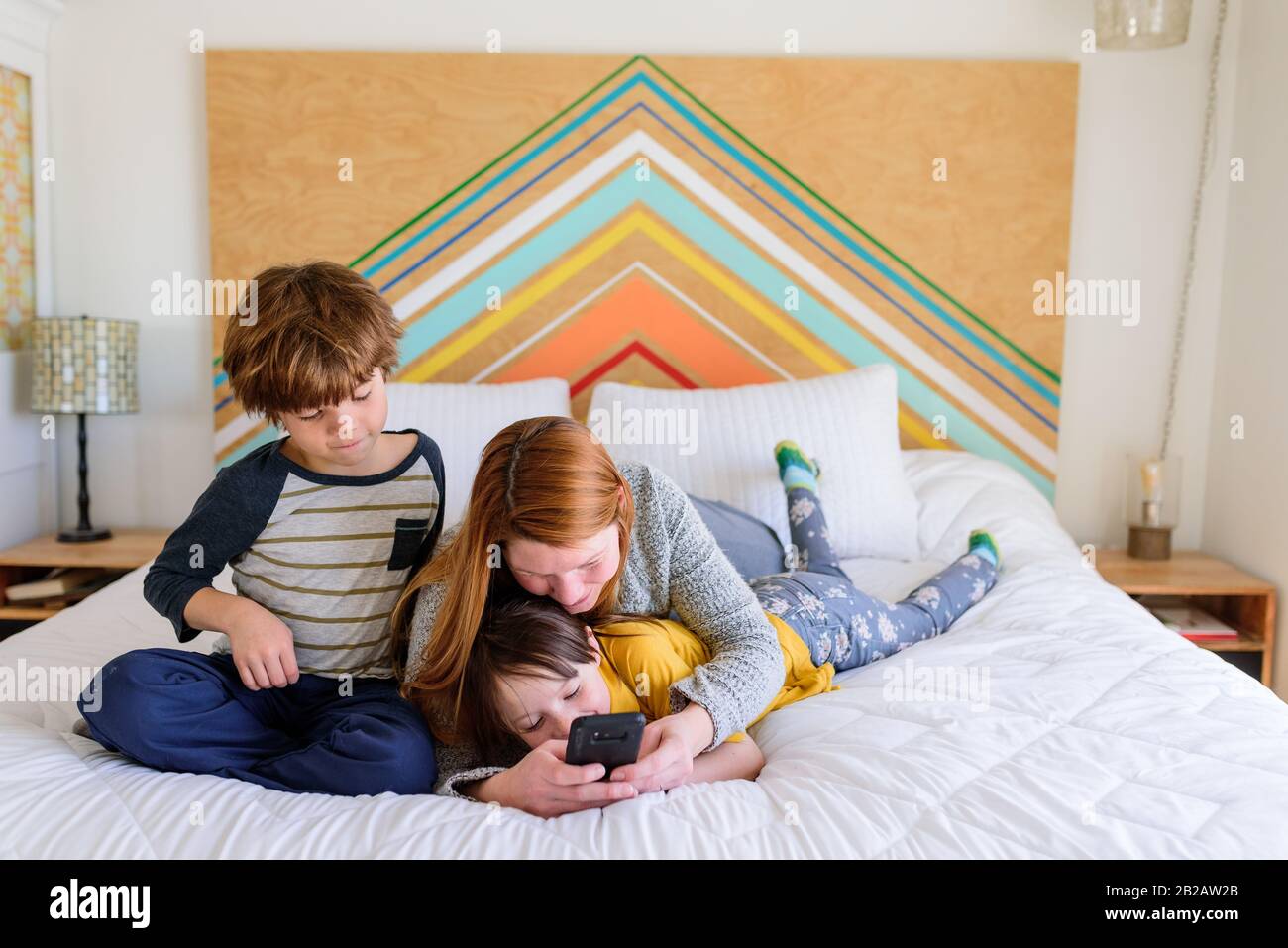 Madre sdraiata sul letto con due bambini che guardano un telefono cellulare Foto Stock