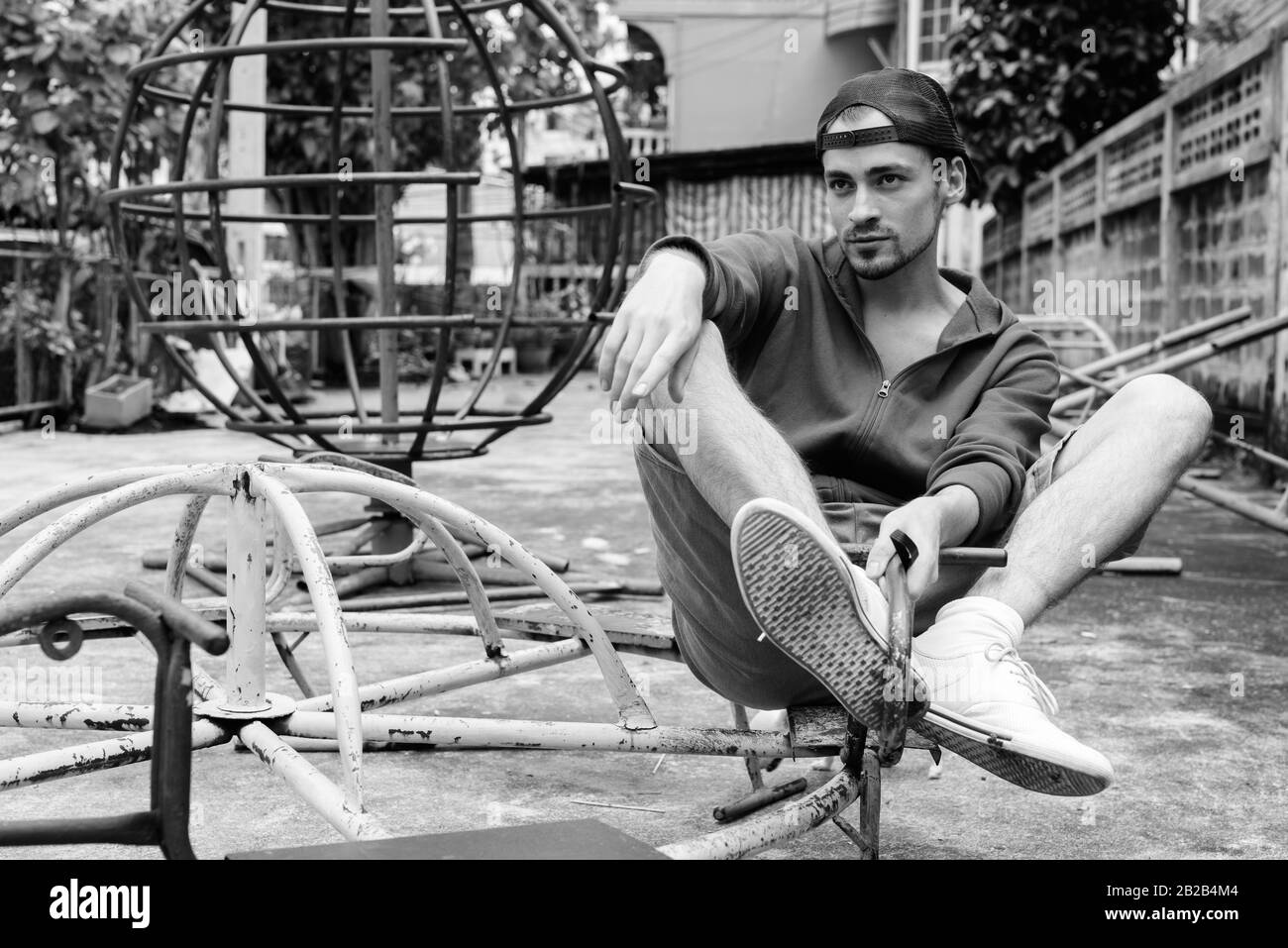 Giovane uomo barbuto pensare mentre è seduto al vecchio arrugginito Merry Go Round e indossando il cappuccio nel vecchio parco giochi Foto Stock