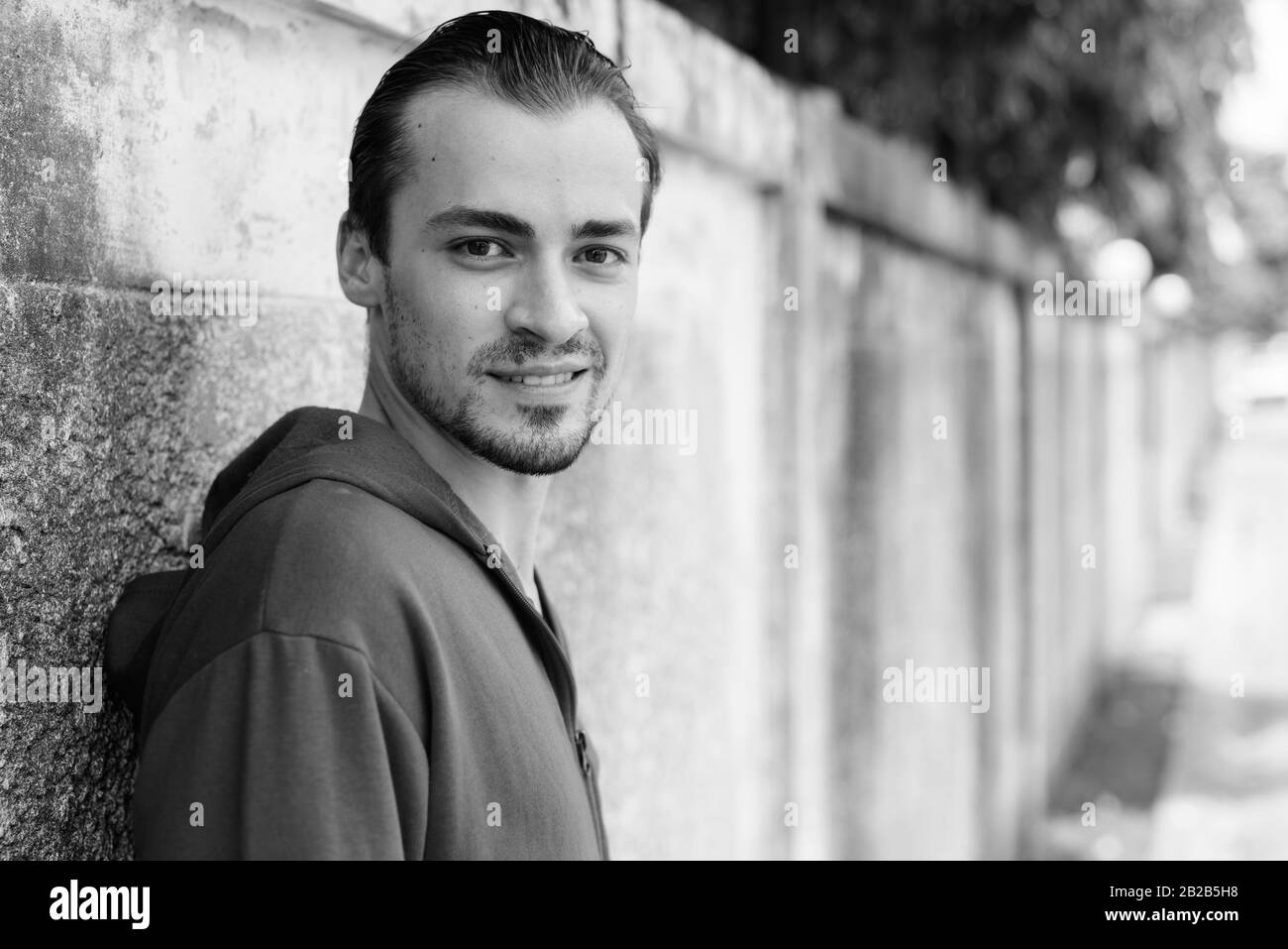 Felice giovane uomo barbuto sorridere mentre indossa felpa con cappuccio rosso e poggiante sul vecchio muro di cemento nelle strade all'aperto Foto Stock