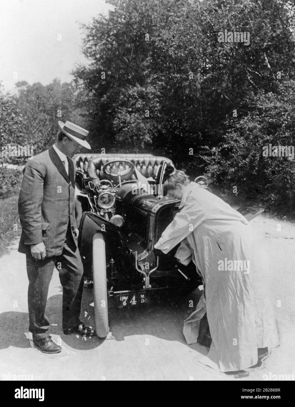 Una donna, che indossa uno smock per evitare di sporcarsi, avvia il motore della sua auto per mezzo di una manovella, mentre l'uomo alla sua sinistra la guarda. Foto Stock