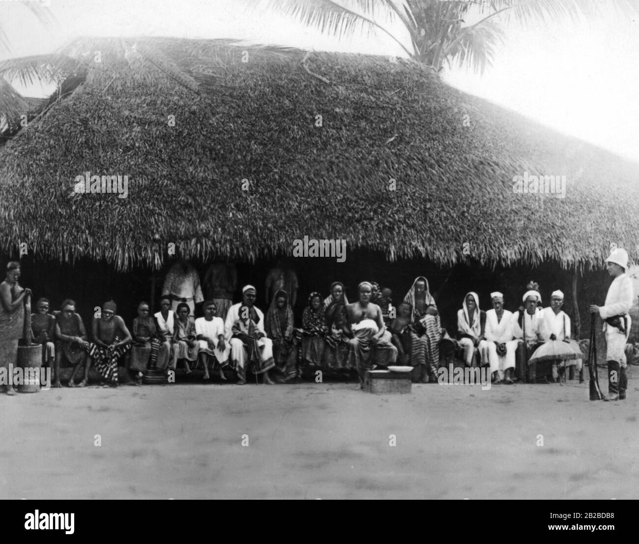 Nativi di fronte alla capanna di un villaggio anziano in Africa orientale tedesca. Lato anteriore destro presumibilmente: Carl Peters. La foto è ondulata. Foto Stock