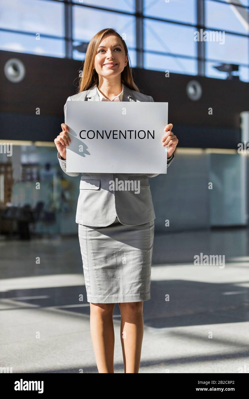 Ritratto di giovane donna d'affari attraente in piedi mentre tiene il bordo bianco con la segnaletica DELLA CONVENZIONE nell'area degli arrivi all'aeroporto Foto Stock