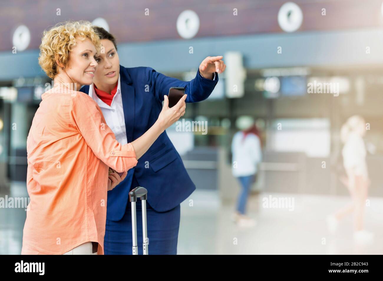 Ritratto di donna matura che mostra il suo smartphone, mentre il personale dell'aeroporto indica la direzione Foto Stock