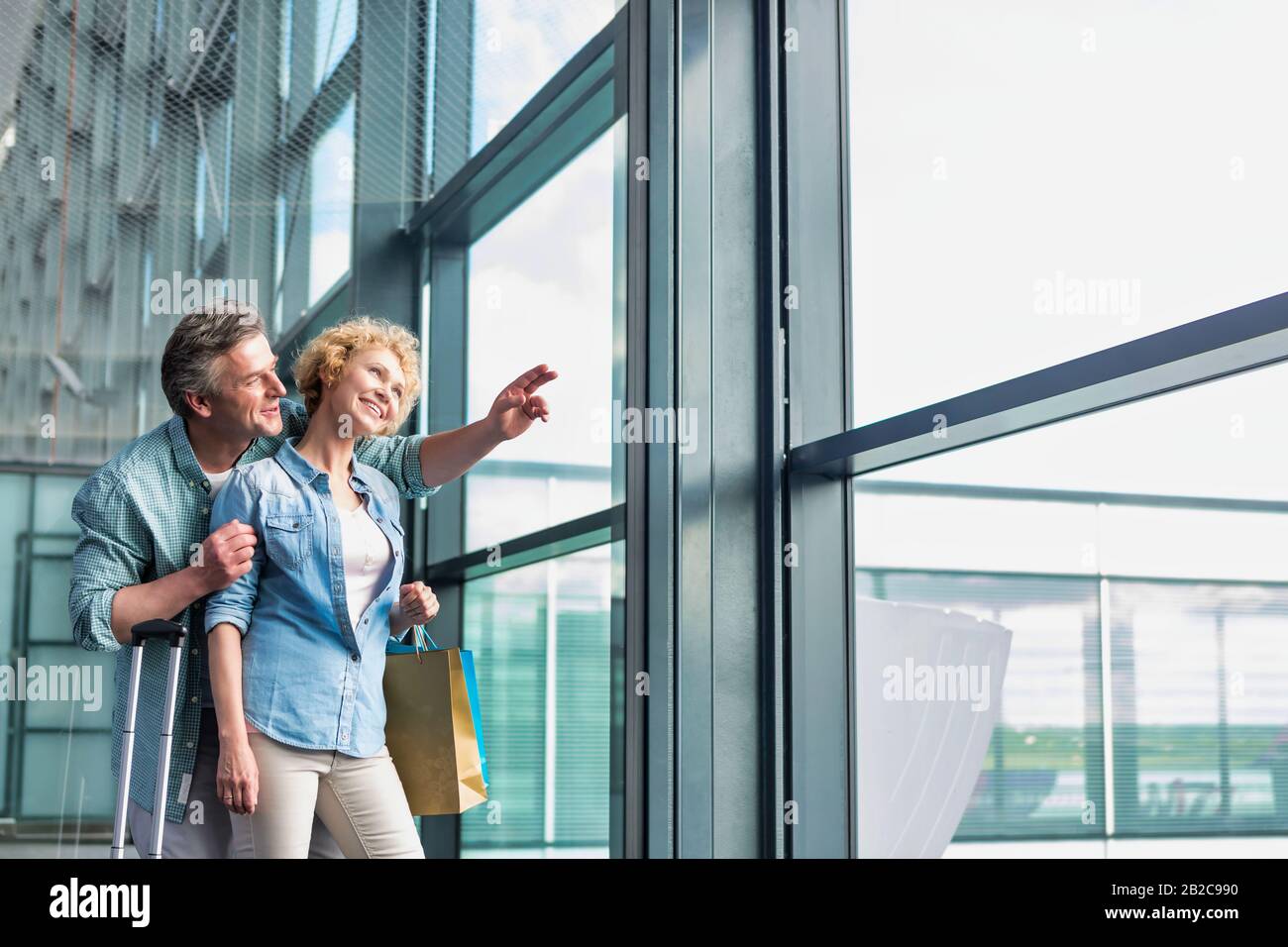 Uomo maturo che abbraccia sua moglie da dietro mentre guarda attraverso la finestra in aeroporto Foto Stock