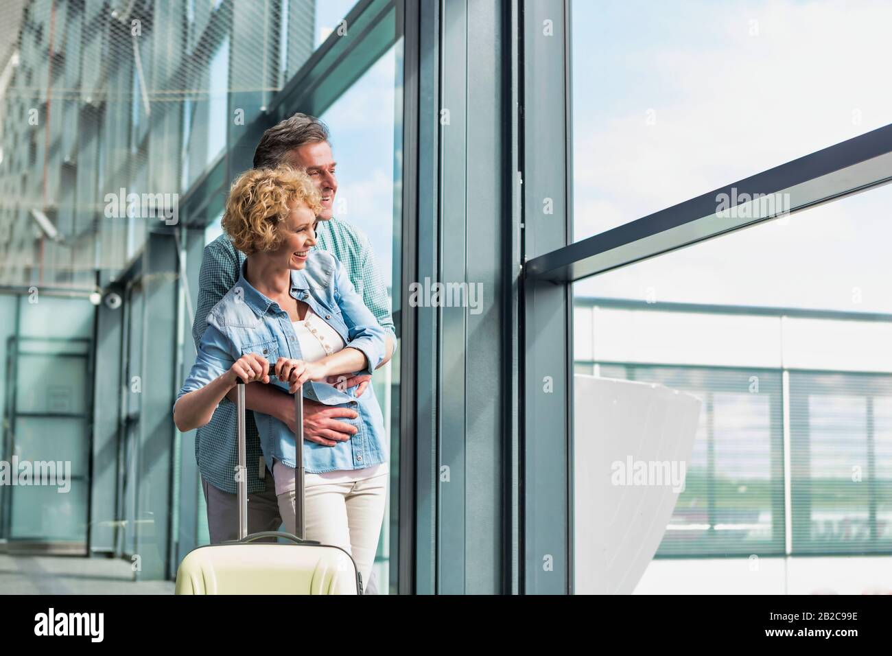 Uomo maturo che abbraccia sua moglie da dietro mentre guarda attraverso la finestra in aeroporto Foto Stock