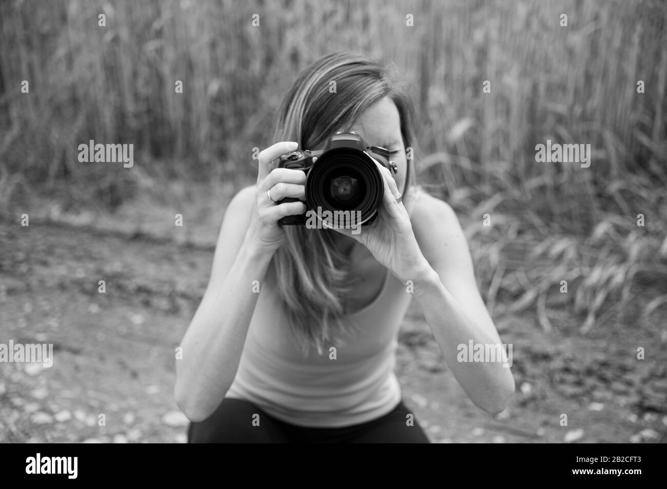 Immagine monocromatica di una giovane fotografa femminile inginocchiata fuori dalla natura scattando una foto direttamente alla fotocamera. Foto Stock