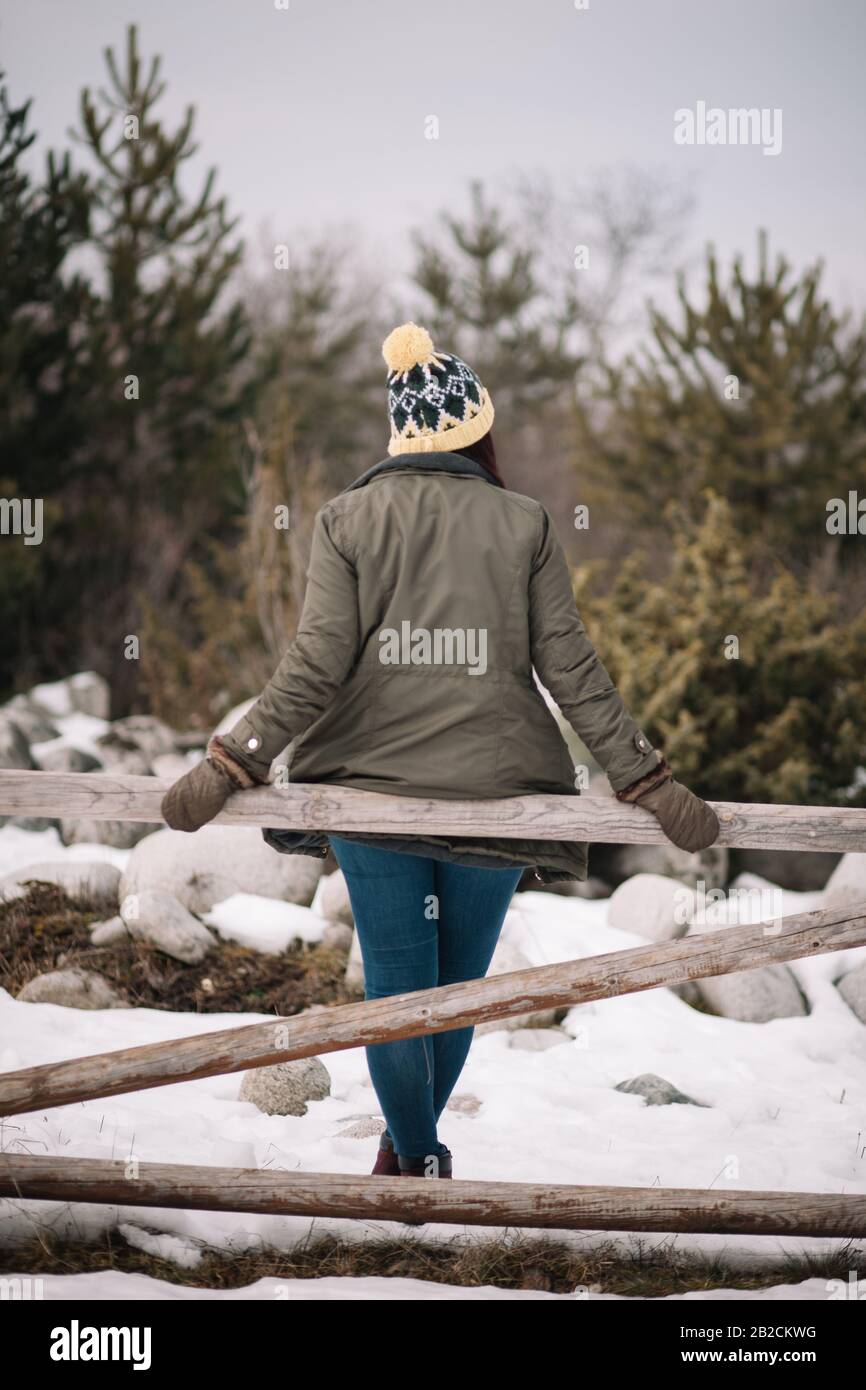 Retro di una donna appoggiarsi sulla recinzione nella foresta. Vista posteriore di una ragazza che indossa cappello e cappotto appoggiato su una recinzione in legno con neve. Foto Stock