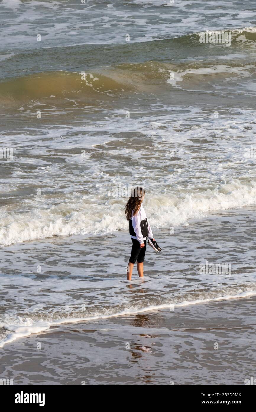 Una giovane donna che pagaia al mare nel surf completamente vestita reggendo le sue scarpe. Foto Stock