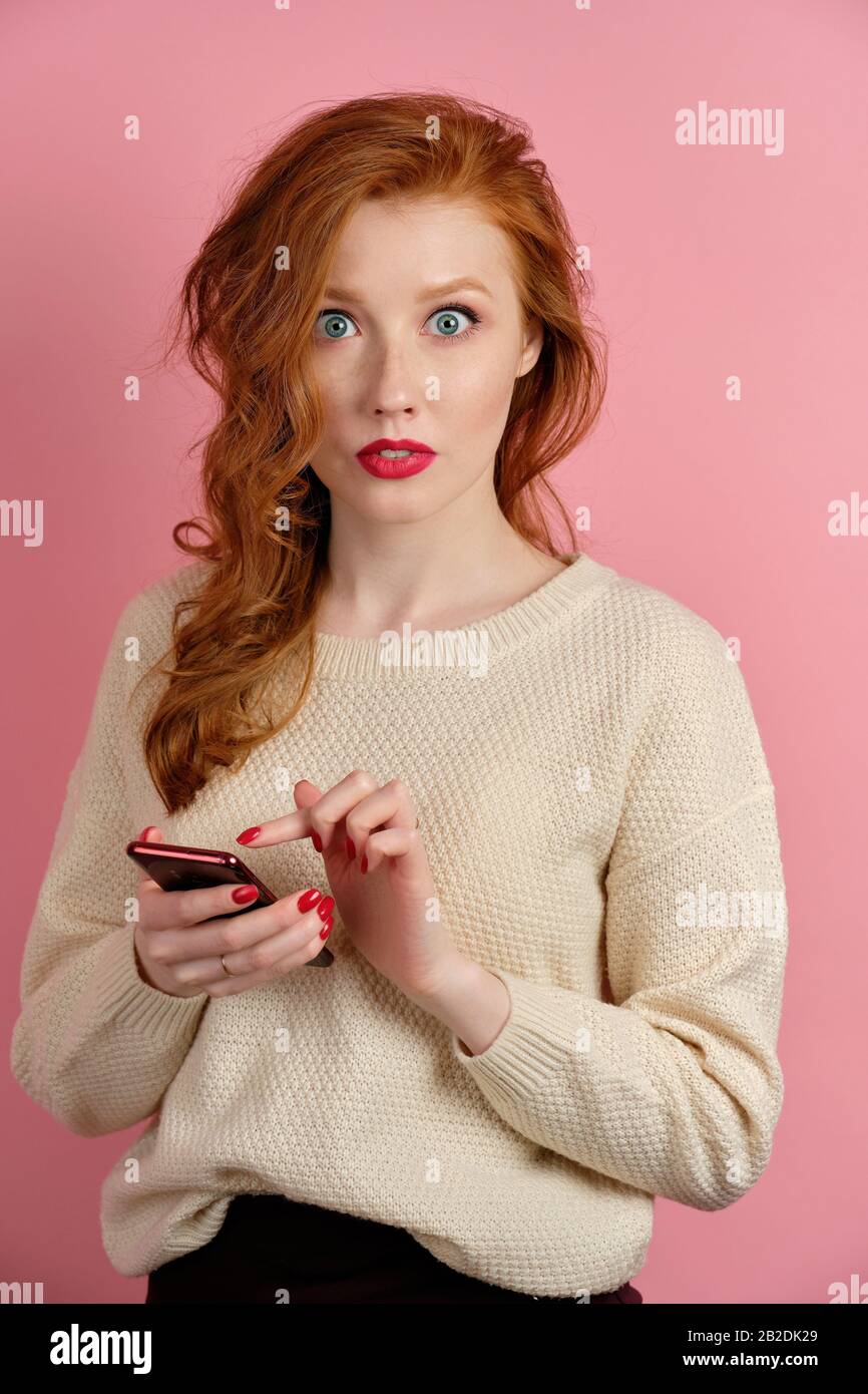 Una ragazza dai capelli rossi con rossetto rosso si alza su uno sfondo rosa  con un telefono in mano e guarda in sorpresa alla fotocamera Foto stock -  Alamy