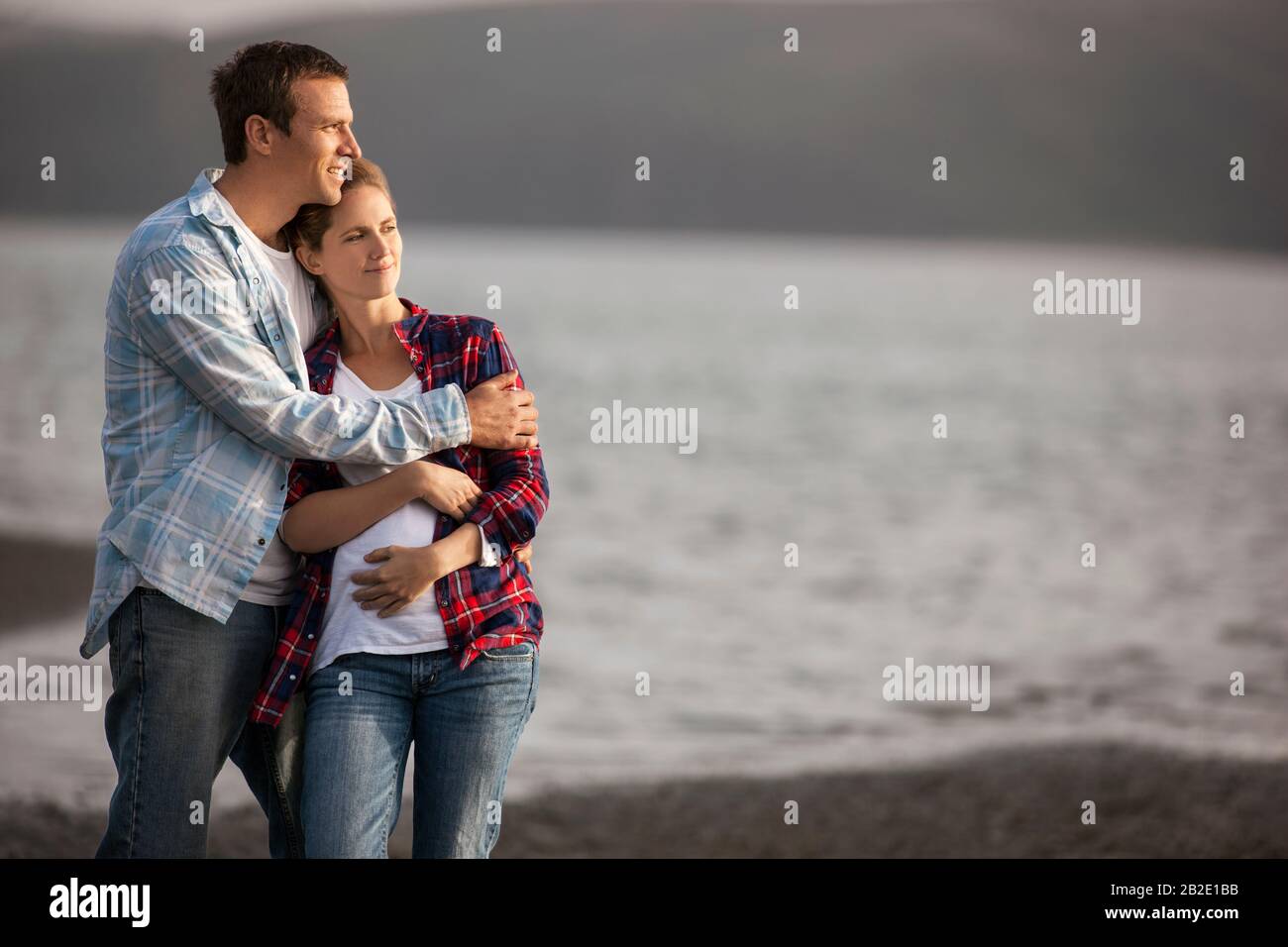 Uomo medio adulto che abbraccia delicatamente la moglie incinta sulla spiaggia Foto Stock