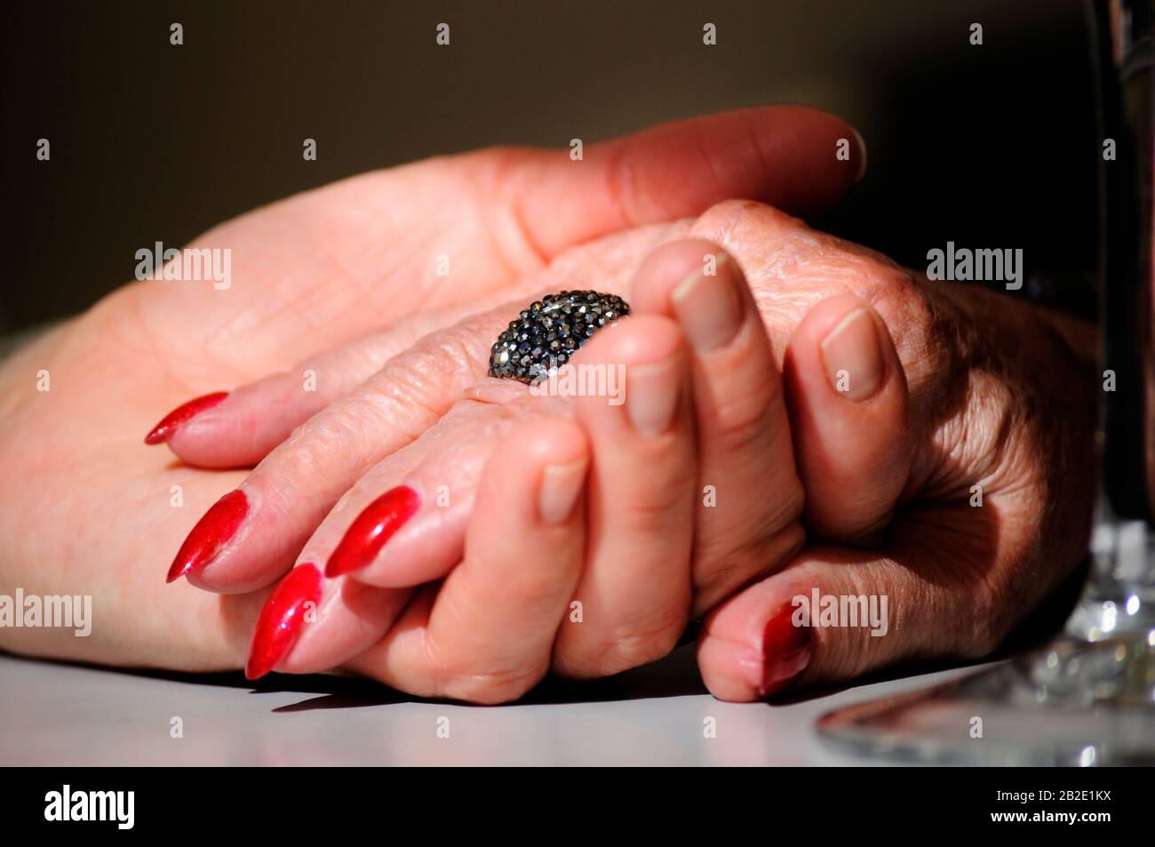 Figlia adulta che tiene la madre stropicciata vecchia mano indossando un anello e smalto rosso del chiodo. Scena parzialmente illuminata con un effetto soft drammatico. Mani sulla tabella Foto Stock