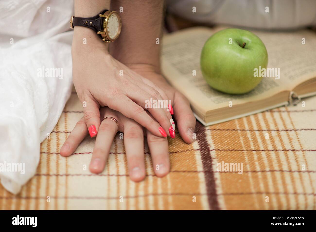 due paia di mani di un uomo e di una donna si attaccano insieme su una leggera copertina morbida e una mela verde e un libro sullo sfondo Foto Stock