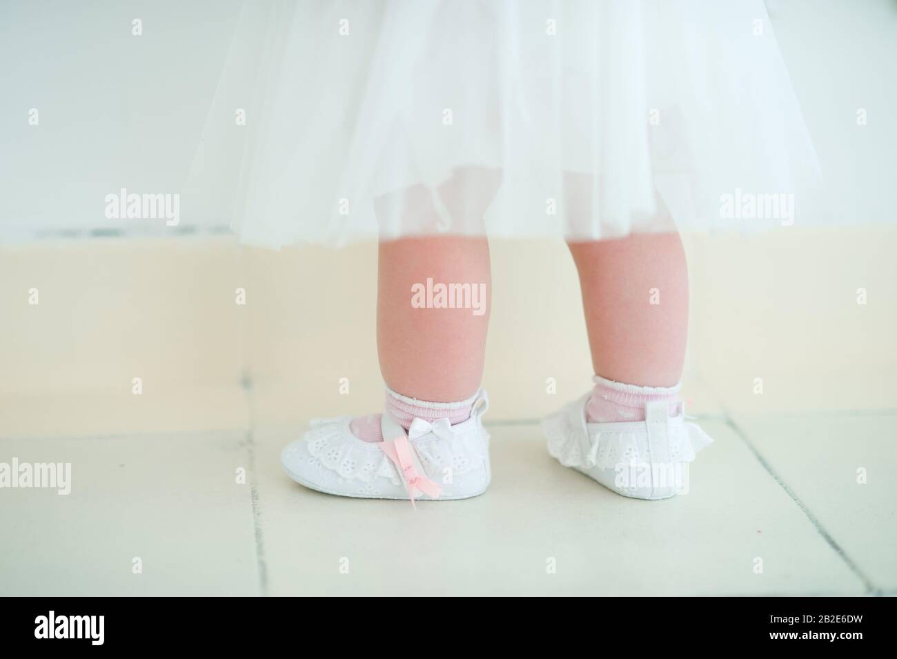 gambe per bambini in scarpe e calzini bianchi su un pavimento di legno chiaro, un morbido abito bianco tulle Foto Stock