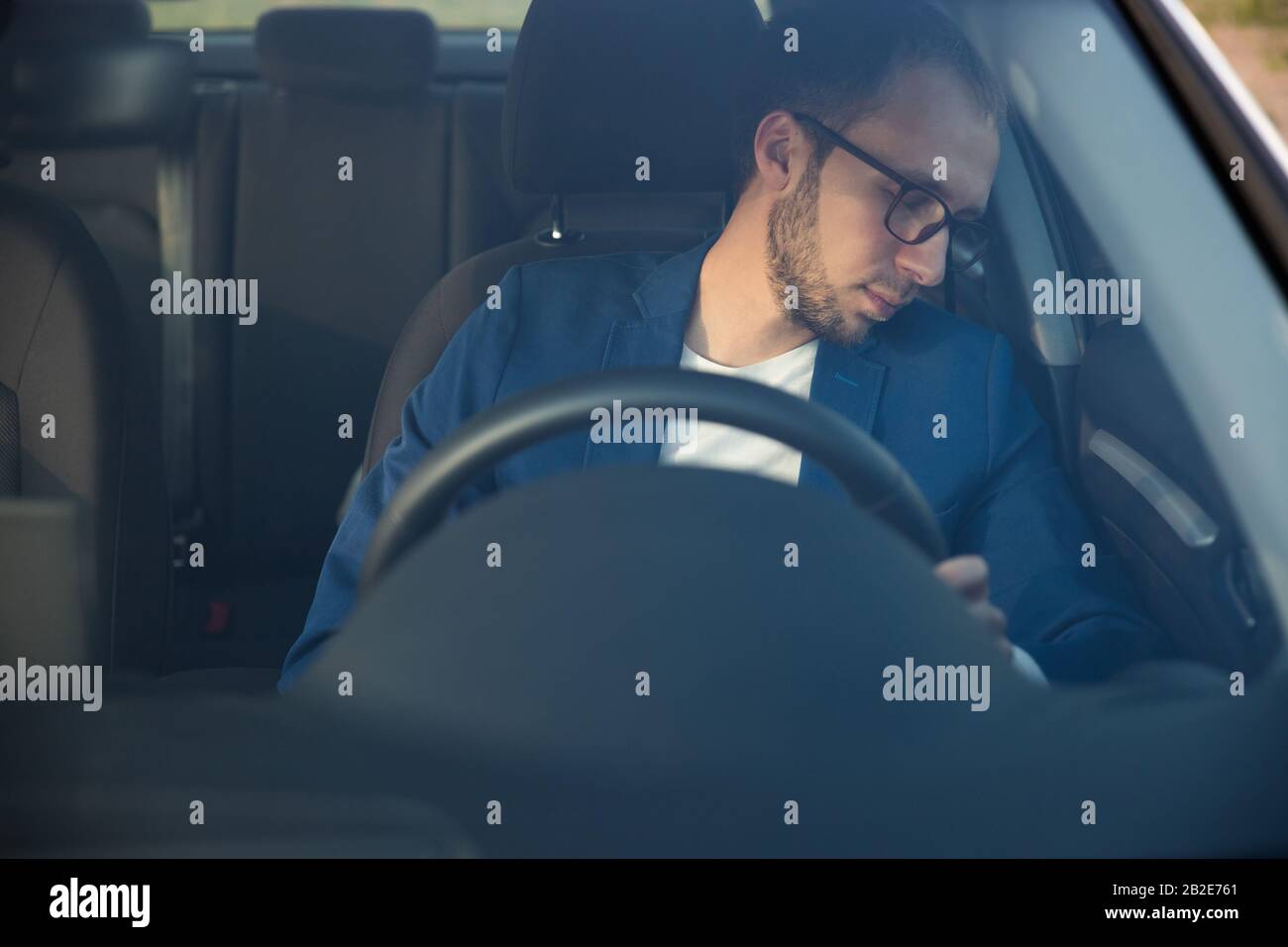 Un uomo d'affari stanco che si addormenta seduto all'interno della sua auto visto attraverso il parabrezza, si è fermato a riposare dopo la guida. Sfinito, sonnolenza, superlavorato dri Foto Stock