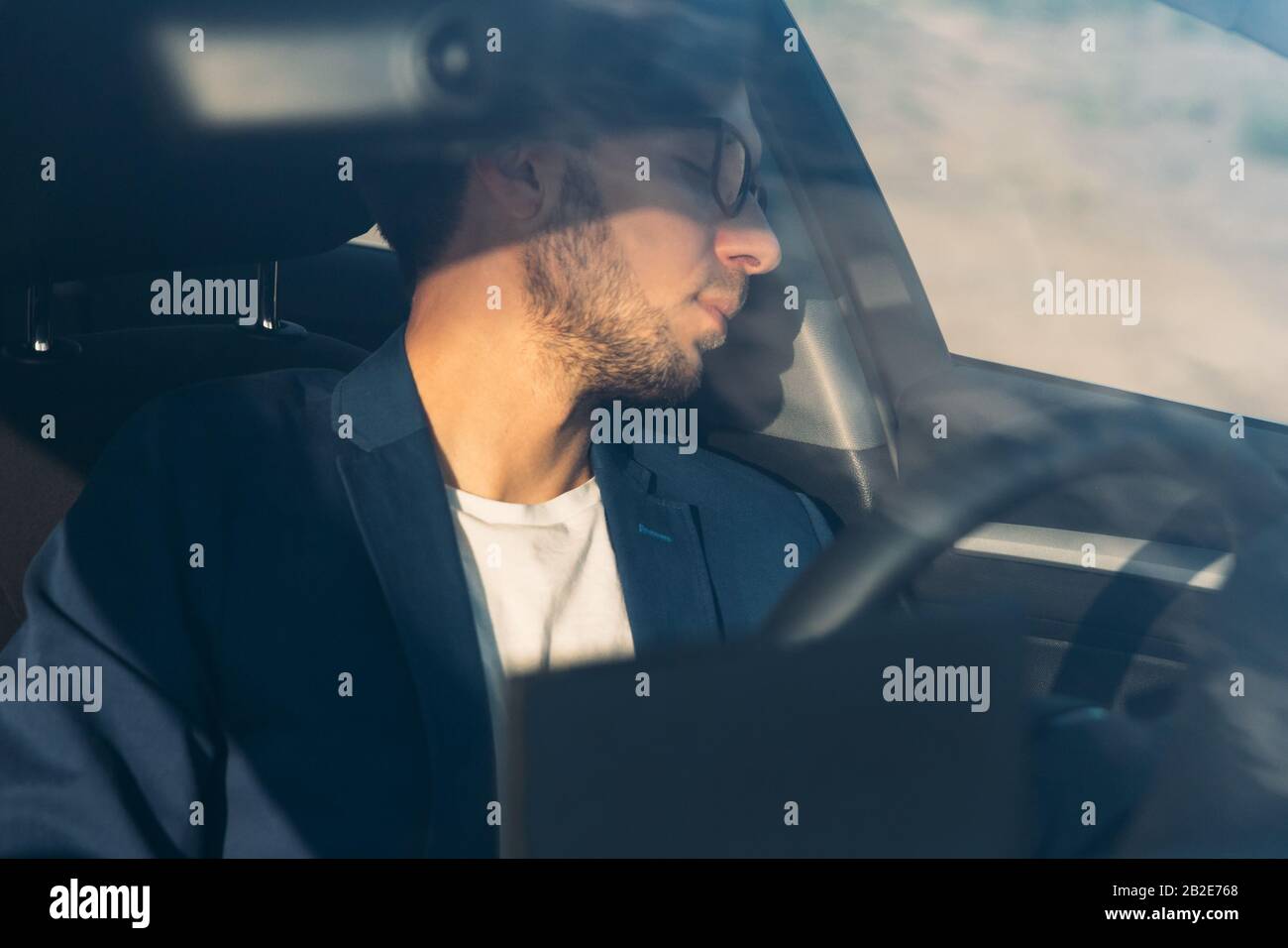 Un uomo d'affari stanco che si addormenta seduto all'interno della sua auto visto attraverso il parabrezza, si è fermato a riposare dopo la guida. Sfinito, sonnolenza, superlavorato dri Foto Stock