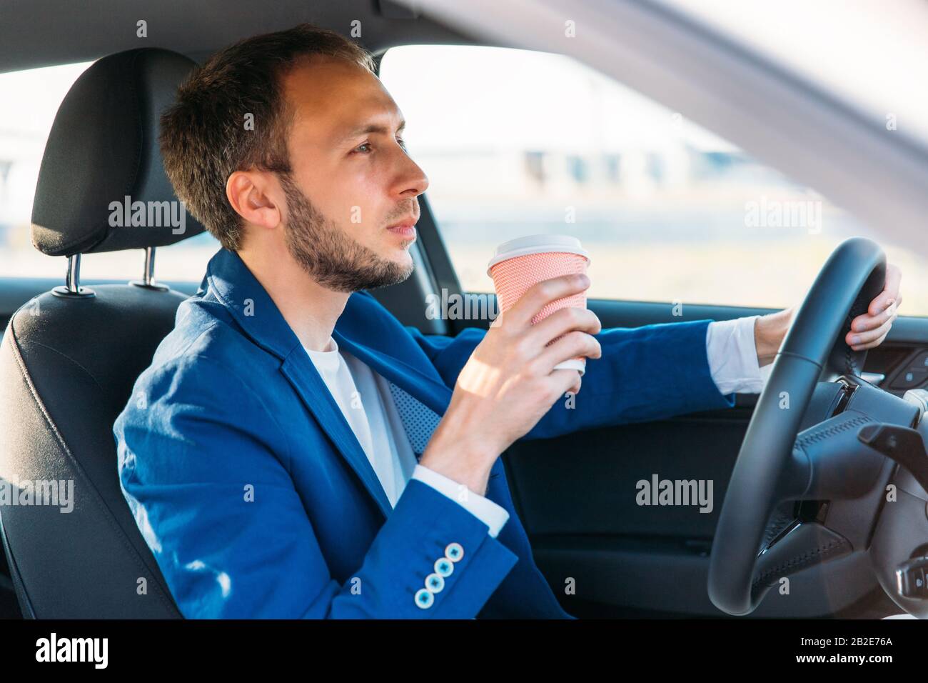 Uomo d'affari in un blazer blu che tiene il volante e beve il caffè da una tazza di carta mentre guida un automobile in modo da non cadere addormentato al volante, lo Foto Stock