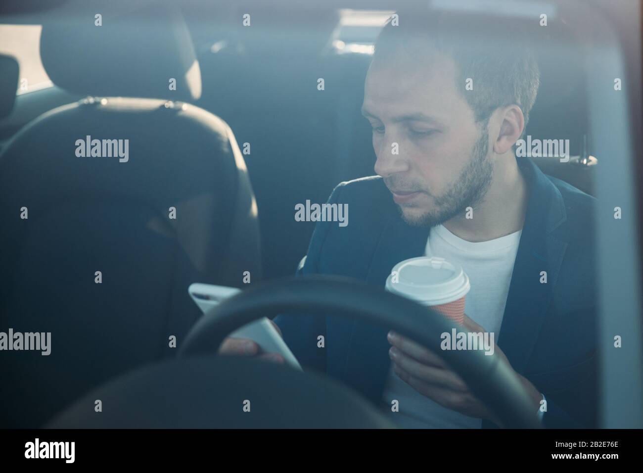 Vista frontale del giovane uomo d'affari seduto in auto e digitando / messaging sullo smartphone, bere caffè in una tazza di carta, visto attraverso il parabrezza. Caffè b Foto Stock
