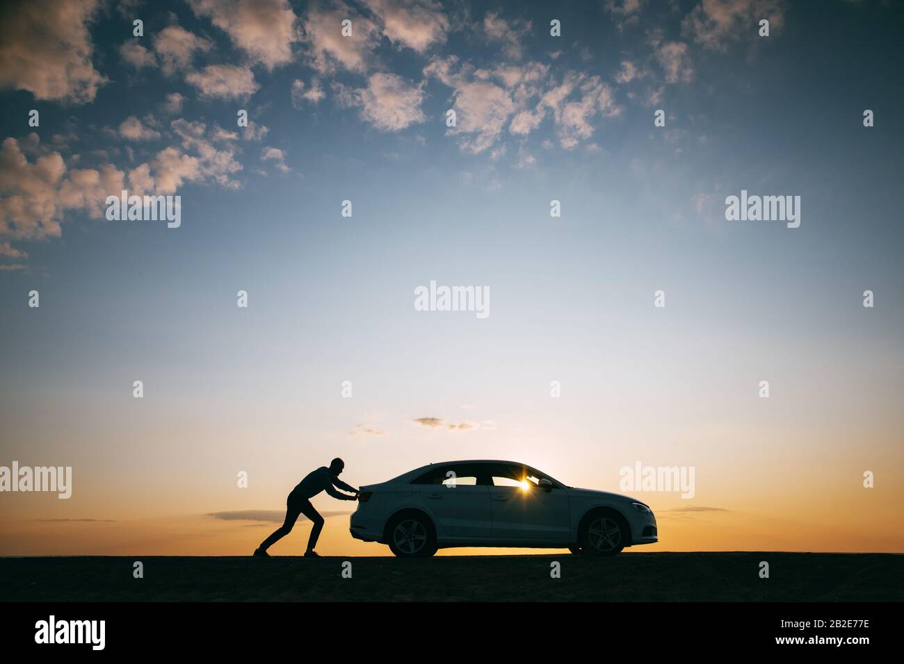 Silhouette di uomo conducente spingere la sua auto lungo una strada vuota dopo la rottura al tramonto, copia spazio, vista laterale. Foto Stock