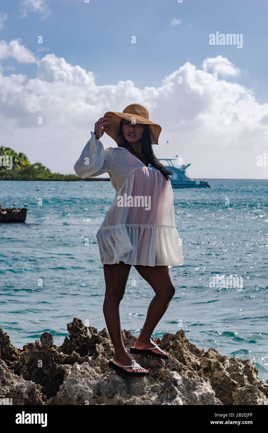 Donna nera da 25 a 30 anni, in piedi su rocce, vestita di modellazione bianca, fresco estate e abiti tropicali, con paesaggio spiaggia e sole s Foto Stock