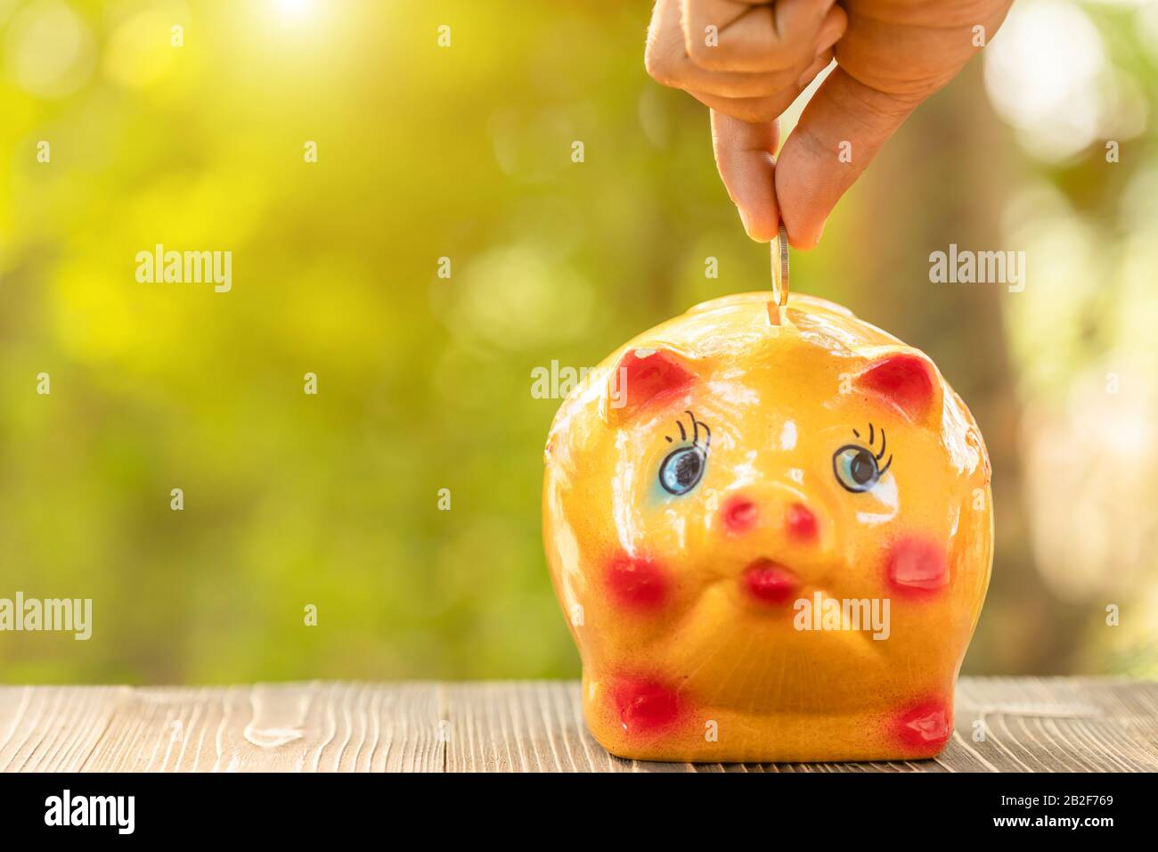 Primo piano mano mettere la moneta a banca piggy con verde natura sfondo sfocato. Concetto di risparmio economico Foto Stock