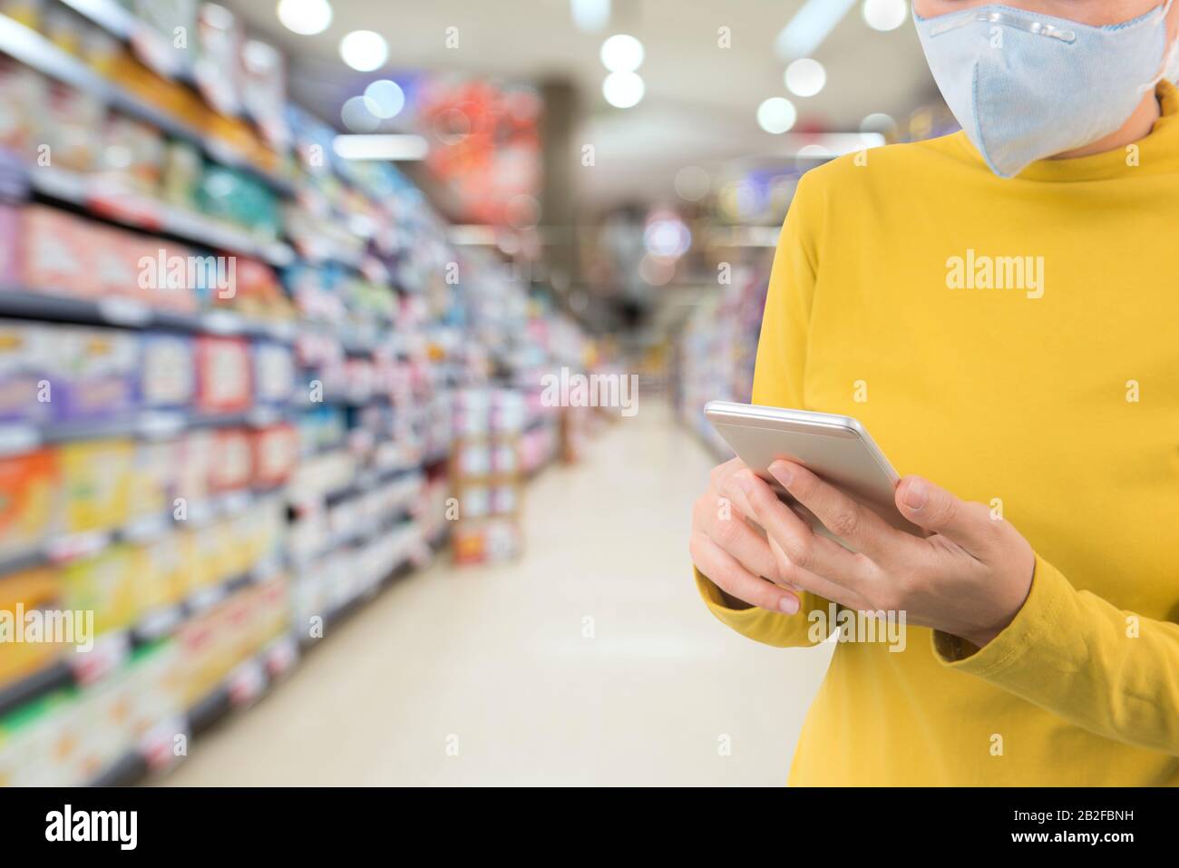 Donna che indossa una maschera protettiva utilizzando il telefono cellulare su uno sfondo supermercato. Proteggersi dalle infezioni virali nella crisi del virus Corona 2020. Foto Stock