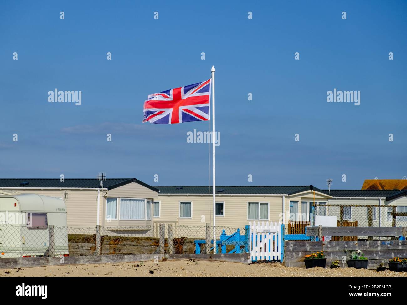 Statico caravan bianco dietro una recinzione con spiaggia di ghiaia e bandiera britannica in pole al West Sands Caravan Park, Bunn Leisure, Selsey, West Sussex, Inghilterra. Foto Stock