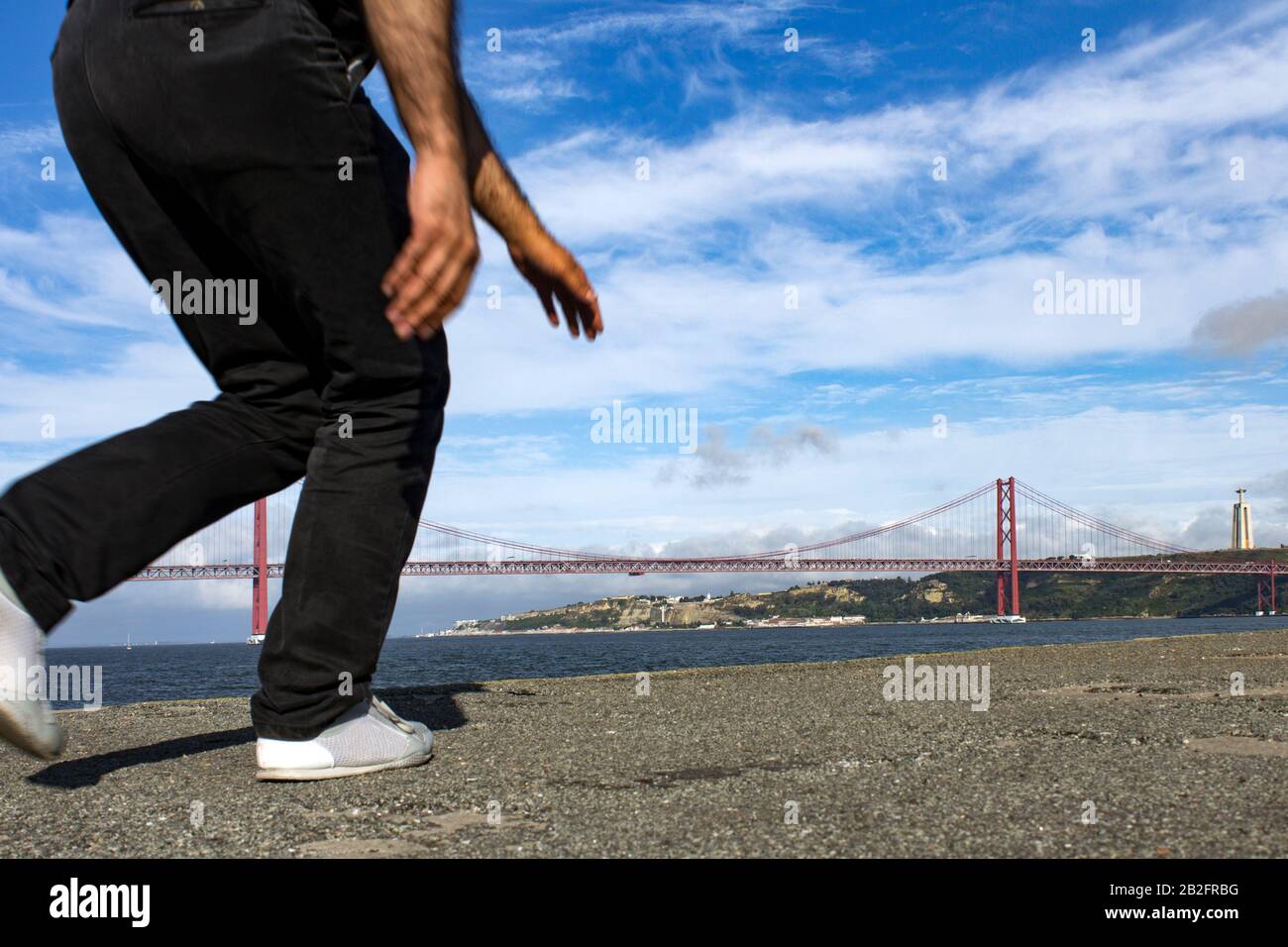 Immagine a basso angolo della metà Inferiore del corpo di un uomo in corsa visto di fronte al ponte 25 de Abril a Lisbona, Portogallo, in una giornata estiva con sk blu Foto Stock