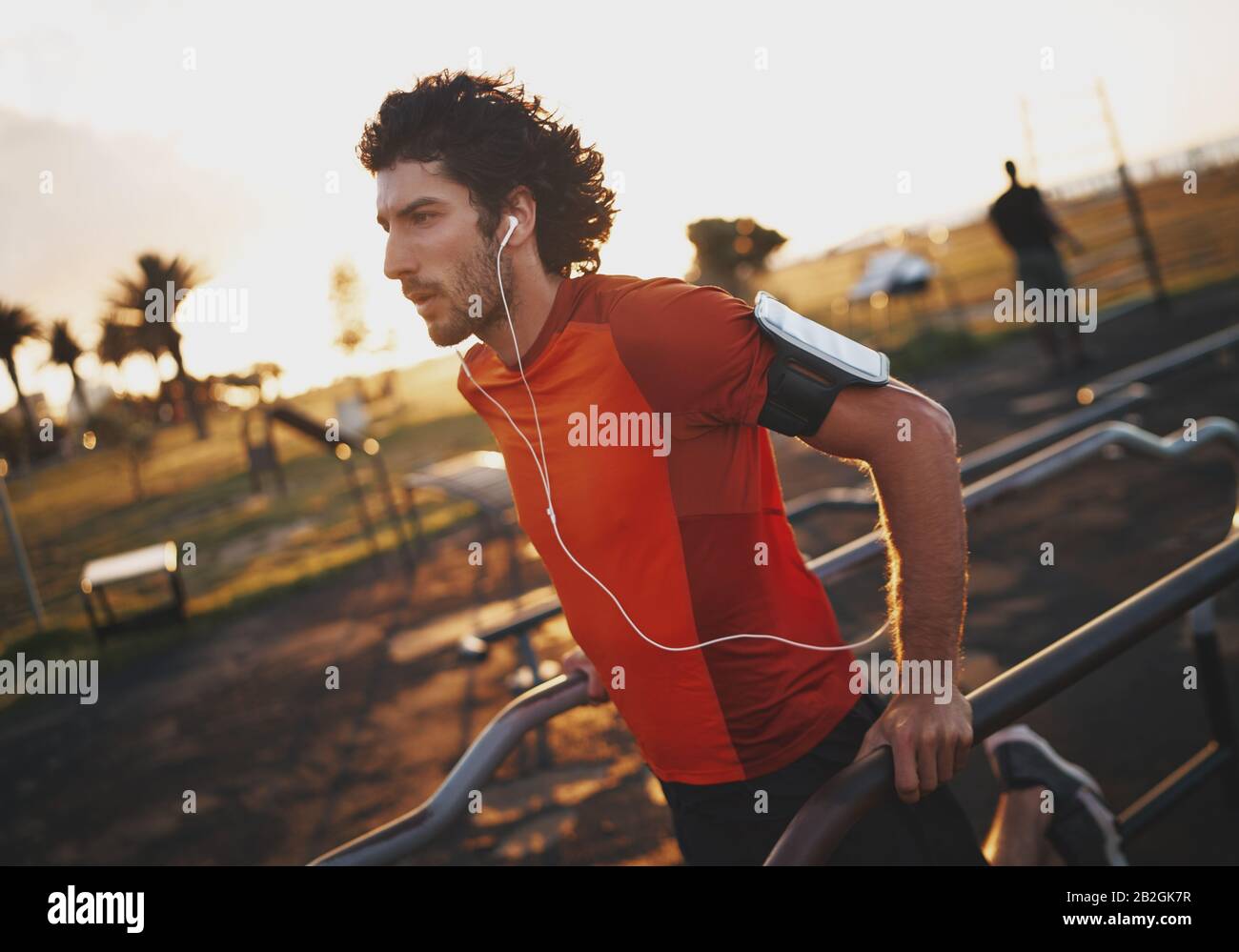 Ritratto di un giovane atletico che ascolta la musica sulle cuffie che si esercitano in un parco fitness all'aperto al mattino - giovane uomo che fa tuffi all'aperto Foto Stock